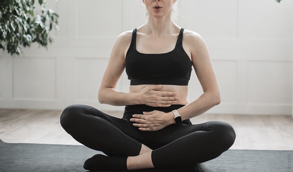 Foto: Eine Person sitzt im Schneidersitz auf einer Matte, hält die Handflächen auf den Bauch und macht Atemübungen